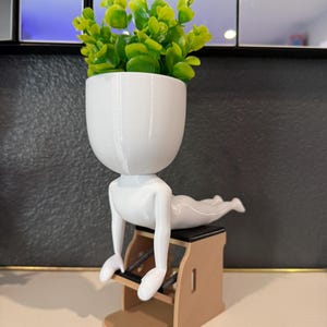 May include: A white planter shaped like a person doing Pilates, with a head-shaped pot filled with green artificial plants. The figure is positioned on a small, tan Pilates chair with a black seat.