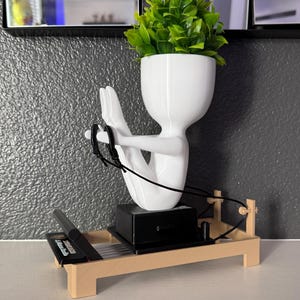 May include: A white abstract human-shaped planter containing a green faux plant, positioned on a beige miniature Pilates reformer machine. The figure is in a seated pose with arms extended, holding black cords attached to the reformer frame.