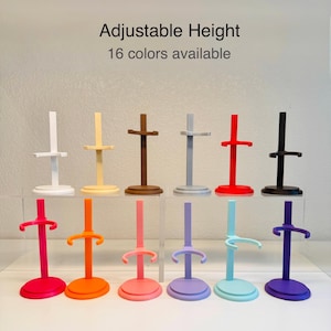 May include: A collection of colorful doll stands in various shades, including white, yellow, brown, gray, red, black, pink, orange, and purple. The stands have an adjustable height feature and are designed to hold dolls. Text on the image reads "Adjustable Height, 16 colors available."