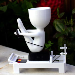 May include: A white 3D-printed planter shaped like a stylized figure performing a rowing exercise on a white machine base. The figure has a hollow head for a plant, a black seat, and black resistance cords. The base features small red and yellow accents.