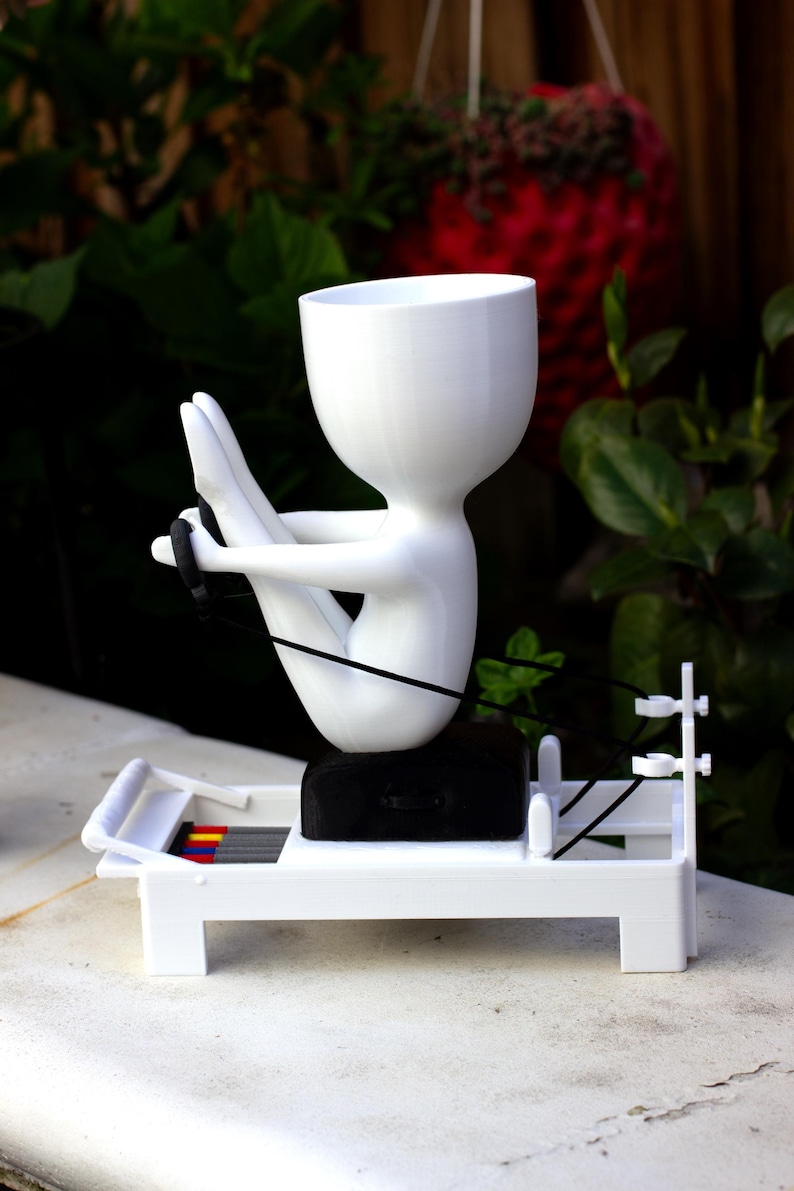 May include: A white 3D-printed planter shaped like a stylized figure performing a rowing exercise on a white machine base. The figure has a hollow head for a plant, a black seat, and black resistance cords. The base features small red and yellow accents.