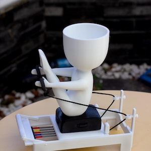 May include: A white planter shaped like a stylized figure sitting on a white Pilates reformer machine. The figure has a round head and holds black resistance bands. The reformer base features a black seat and a set of small red, yellow, and blue rods.