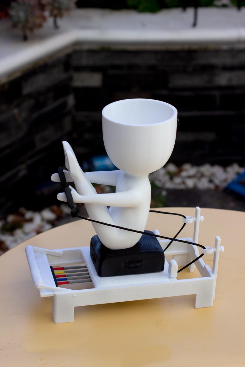 May include: A white planter shaped like a stylized figure sitting on a white Pilates reformer machine. The figure has a round head and holds black resistance bands. The reformer base features a black seat and a set of small red, yellow, and blue rods.