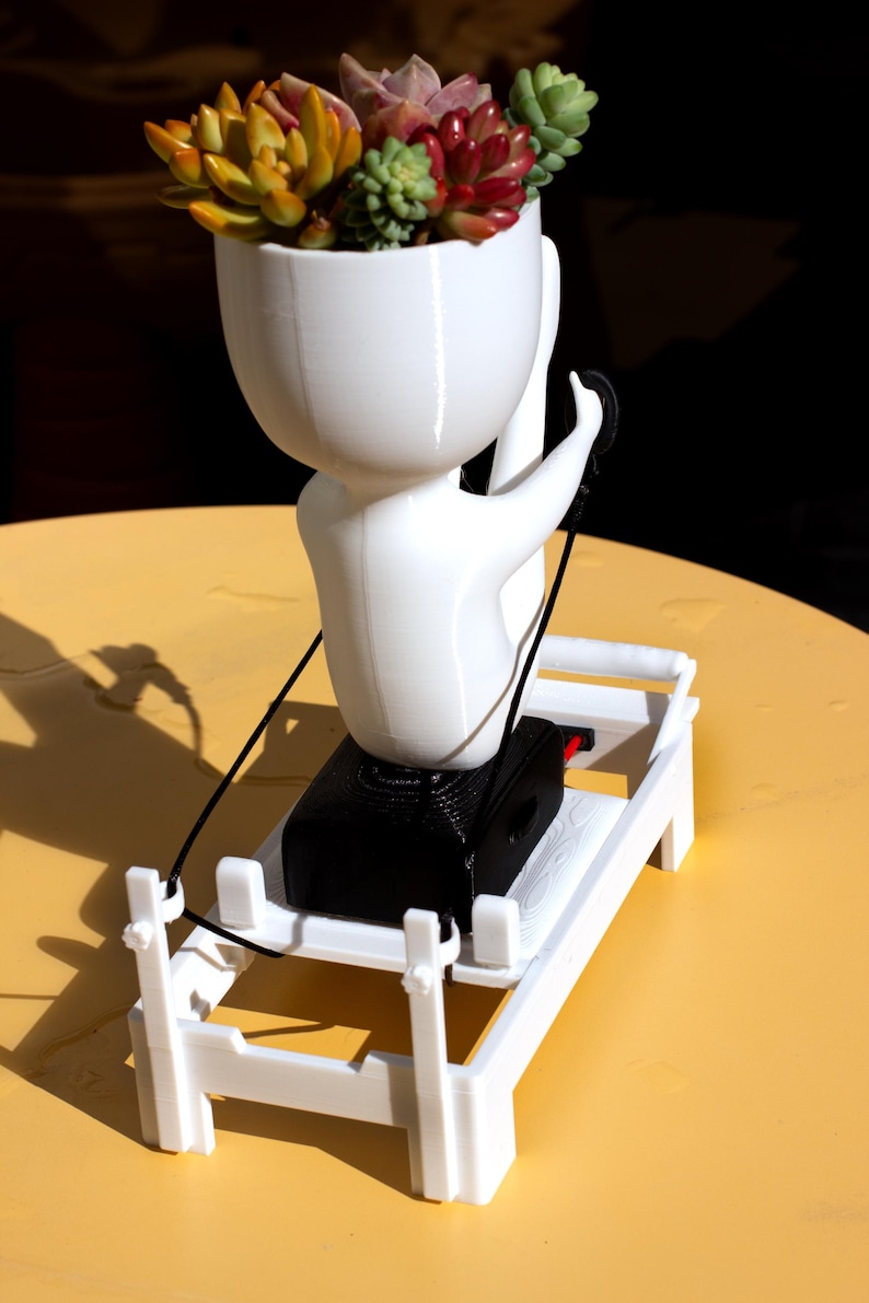 May include: A white 3D-printed planter shaped like a stylized figure performing a yoga pose, filled with colorful succulents. The figure sits on a black base attached to a white rectangular frame with a thin black cord extending from the figure.