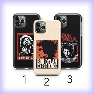 May include: Three different phone cases featuring Bob Dylan. The first case is black with a red and white image of Bob Dylan singing. The second case is beige with a red and white image of Bob Dylan's face. The third case is black with a red and white image of Bob Dylan in concert. The text on the third case reads "Bob Dylan in Concert Summer Tour and His Band".