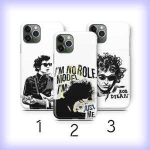 May include: Three white iPhone cases with different designs featuring a black and white illustration of Bob Dylan. The first case shows Dylan playing a guitar. The second case shows Dylan with the text "I'm no role model I'm just me". The third case shows Dylan's face with the text "Bob Dylan".