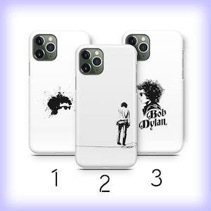 May include: Three white iPhone cases with different designs. The first case has a black ink splatter design. The second case has a black and white silhouette of Bob Dylan. The third case has a black and white silhouette of Bob Dylan with the text "Bob Dylan" in black.