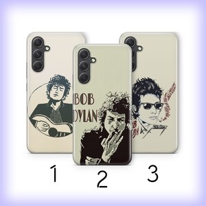 May include: Three phone cases featuring different images of Bob Dylan. The first case shows a black and white image of Bob Dylan playing a guitar. The second case shows a black and white image of Bob Dylan smoking a cigarette. The third case shows a black and white image of Bob Dylan with sunglasses. All three cases have a beige background with the text "Bob Dylan" in red and black.