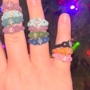 May include: A hand displays several macrame rings in various colors, including pink, teal, green, and blue. Each ring features a central stone and gold-colored beads. The rings are handmade and showcase intricate knotting patterns, perfect for adding a bohemian touch.