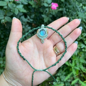 May include: A handmade macrame bracelet with a central pendant. The pendant features a round, iridescent blue gemstone, surrounded by a light green macrame design and small gold beads. The bracelet is made of dark green cord and has an adjustable closure.