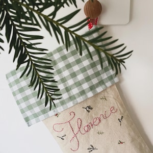 May include: A festive Christmas stocking featuring a green and white gingham cuff. The name "Florence" is embroidered in red script on the natural-colored fabric. Decorated with embroidered leaves and berries.
