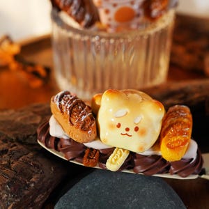 May include: A hair clip featuring a miniature bear-shaped bread loaf, two small brown bread rolls, and dark brown chocolate-colored swirls. The bear face has small eyes, a nose, and a mouth, with a tiny light-colored rectangular piece attached below.