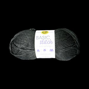 May include: A skein of dark charcoal grey yarn wrapped in a white paper label. The label features the text BASIC stitch anti-pilling and includes small icons of a hat, a scarf, and a sweater. The yarn has a visible twisted fibre texture.
