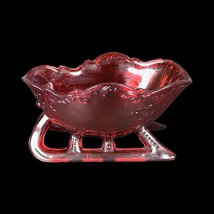 Puede incluir: Un cuenco de cristal rojo translúcido con forma de trineo, que presenta un ornamentado patrón floral en relieve a lo largo del borde superior y una textura de cuentas decorativa en los patines curvos que sostienen la base.