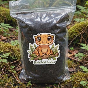 May include: A clear plastic resealable bag filled with dark brown soil substrate. A sticker on the front features a cartoon orange gecko sitting on a log with green leaves and the text Shield Wall Exotics printed in white at the bottom.