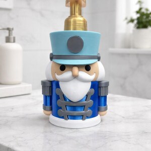 May include: A novelty soap dispenser shaped like a toy soldier nutcracker. The dispenser features a blue hat and uniform with gray and white accents. The pump is gold-colored. The dispenser is on a white countertop.