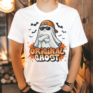 May include: White t-shirt with a cartoon ghost wearing sunglasses and an orange cap. The ghost has its arms crossed, with bats around it. The words "ORIGINAL GHOST" are printed in orange and white on the shirt.