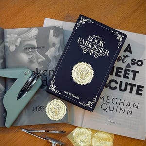 May include: A navy blue book embosser kit with gold foil seals, a mint green handheld embosser, and a book. The book cover reads "BOOK EMBOSSER" with decorative flourishes. The kit includes a book, seals, and tools.