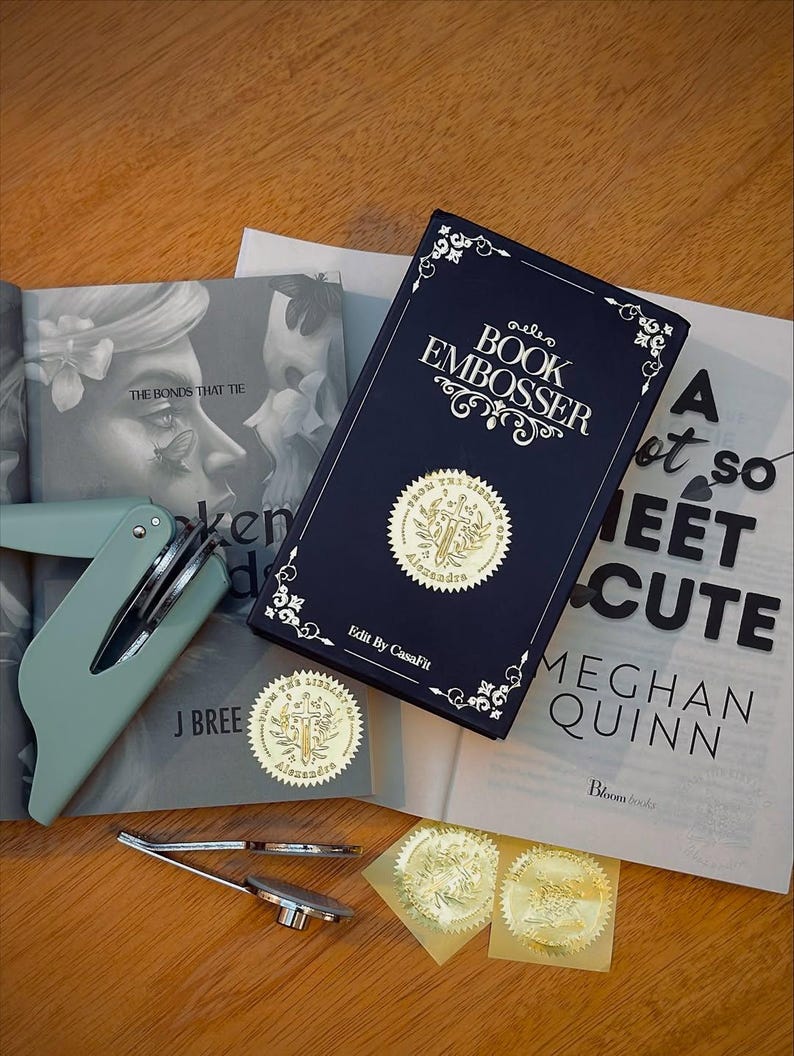 May include: A navy blue book embosser kit with gold foil seals, a mint green handheld embosser, and a book. The book cover reads "BOOK EMBOSSER" with decorative flourishes. The kit includes a book, seals, and tools.