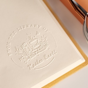 May include: Close-up of a cream-colored book page with a debossed circular design. The design includes the text "FROM THE LIBRARY OF Kailee Scott" surrounding a graphic of a book, bowl, and foliage. A copper-colored object is visible in the background.