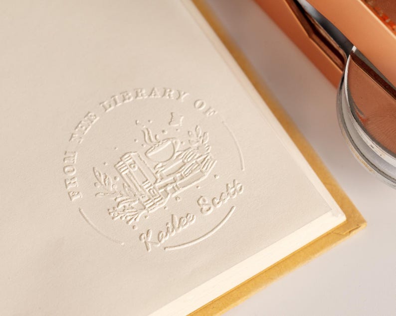 May include: Close-up of a cream-colored book page with embossed text and a circular emblem. The text reads "FROM THE LIBRARY OF Kailee Scott". The emblem features a stack of books, a bowl, and floral elements.