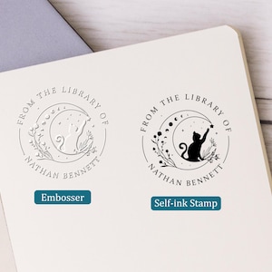 May include: Two circular stamps with a cat reaching for a star inside a crescent moon, surrounded by floral elements. The text "FROM THE LIBRARY OF NATHAN BENNETT" is arched around the design. One stamp is an embosser, the other is a self-ink stamp.