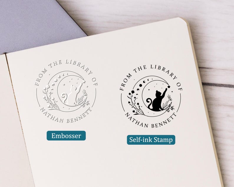 May include: Two circular stamps with a cat reaching for a star inside a crescent moon, surrounded by floral elements. The text "FROM THE LIBRARY OF NATHAN BENNETT" is arched around the design. One stamp is an embosser, the other is a self-ink stamp.