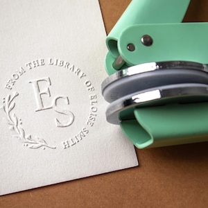 May include: A mint green embosser tool next to a white card with a circular embossed design. The design includes the text "FROM THE LIBRARY OF ELOISE SMITH" around the initials "FS" and a floral element.