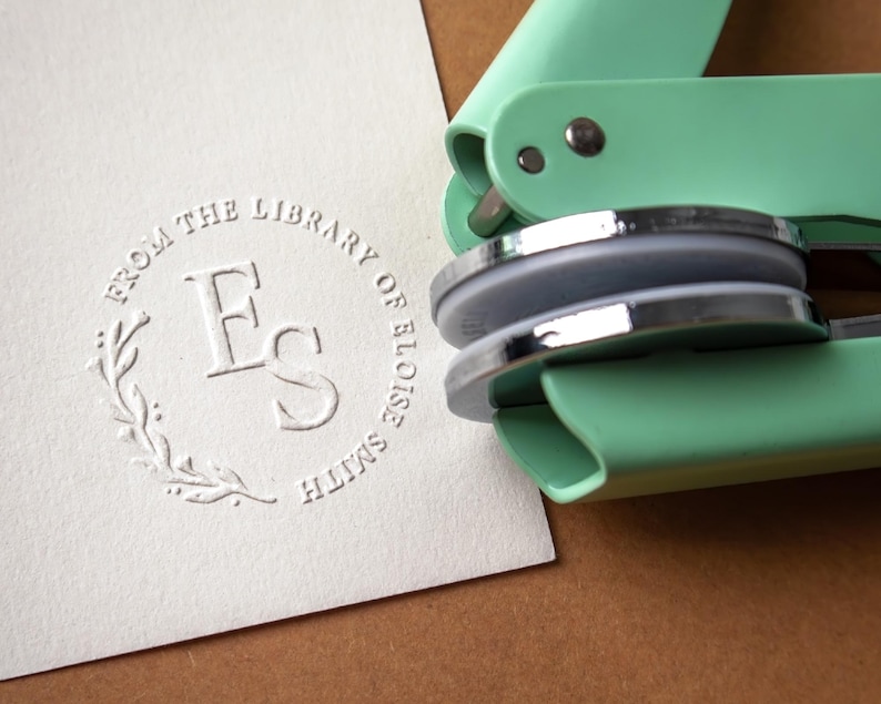 May include: A mint green embosser tool next to a white card with a circular embossed design. The design includes the text "FROM THE LIBRARY OF ELOISE SMITH" around the initials "FS" and a floral element.