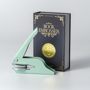 May include: A mint green book embosser next to a black book with gold lettering and a gold seal. The book reads "BOOK EMBOSSER". The embosser has a lever and two circular dies.