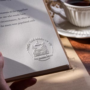 May include: A book with a white page featuring the embossed text "FROM THE LIBRARY OF Benjamin Grant" and a decorative emblem. A white teacup and saucer are in the background. The book is open, with text visible on the page.