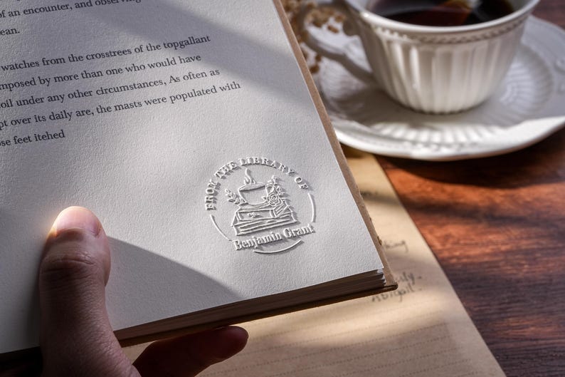 May include: A book with a white page featuring the embossed text "FROM THE LIBRARY OF Benjamin Grant" and a decorative emblem. A white teacup and saucer are in the background. The book is open, with text visible on the page.