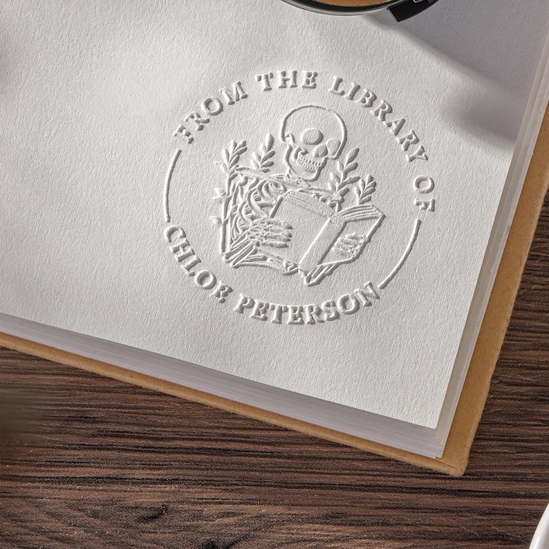 May include: A white embossed bookplate with the text "FROM THE LIBRARY OF CHLOE PETERSON" encircling a skeleton reading a book. The bookplate is on a white page with a tan border, set against a dark wood surface.