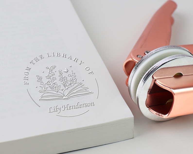 May include: A rose gold book embosser next to a white book with a debossed design. The design reads "From the Library of" with an open book, flowers, and the name "Lily Henderson".