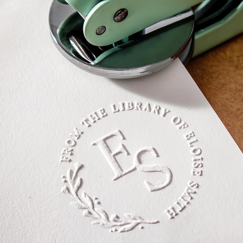 May include: A mint green and silver embossing seal presses a custom design onto a white paper. The embossed design reads "FROM THE LIBRARY OF ELOISE SMITH" around a central "Es" monogram, encircled by a laurel wreath.
