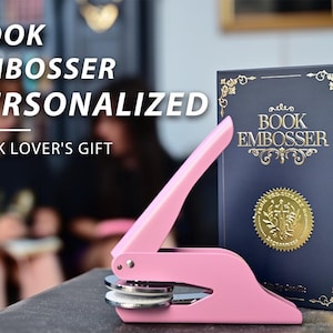 May include: A pink book embosser with a matching book. The book is navy blue with gold lettering and a gold seal. The text on the book reads "BOOK EMBOSSER". The image also includes the text "BOOK EMBOSSER PERSONALIZED BOOK LOVER'S GIFT".