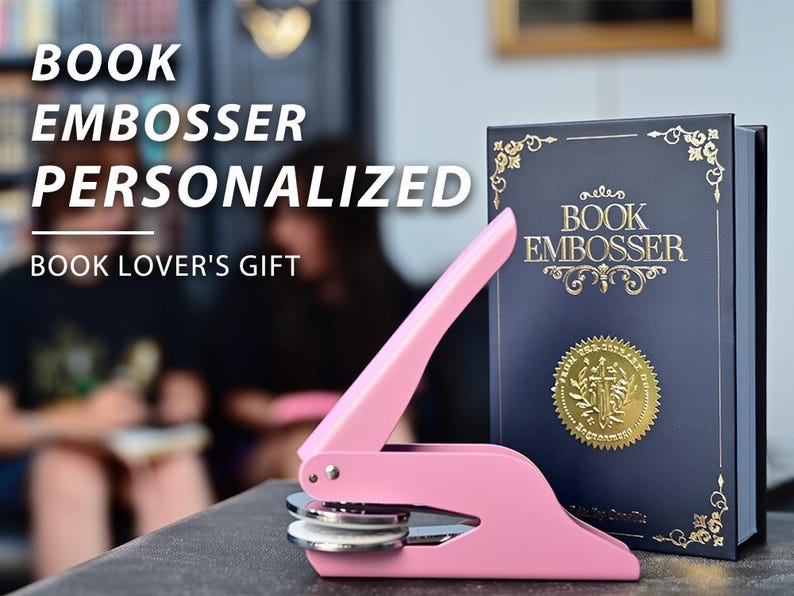 May include: A pink book embosser with a matching book. The book is navy blue with gold lettering and a gold seal. The text on the book reads "BOOK EMBOSSER". The image also includes the text "BOOK EMBOSSER PERSONALIZED BOOK LOVER'S GIFT".