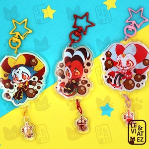 Puede incluir: Tres abalorios de personajes coloridos con abalorios de té de burbujas adjuntos. Cada abalorio presenta un personaje diferente con detalles rojos, negros y blancos. Los abalorios están unidos a una cadena plateada con un cierre en forma de estrella. El texto "LE & VIAT MEZ" es visible en los abalorios.