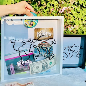 May include: White shadow box with a black wire world map design. The box contains various travel souvenirs, including a Knott's Berry Farm ticket, a Voyage to the Iron Reef ticket, a $1 bill, and a pink and white ticket with the text "Knott's Berry Farm for the Cu."