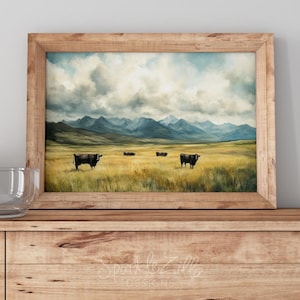 May include: A framed landscape painting depicts a grassy field with several cows grazing. Mountains are in the background under a cloudy sky. The artwork is in a wooden frame and rests on a wooden cabinet.