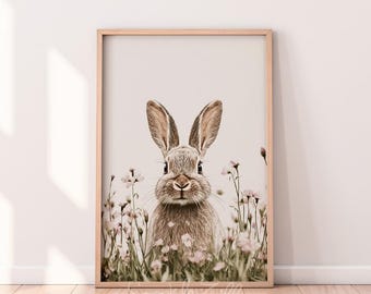 May include: A framed print featuring a close-up of a rabbit in a field of flowers. The rabbit has long ears and a soft, brown and gray coat. The flowers are small and pink, with green stems and leaves. The print is in a light brown wooden frame.