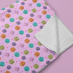 May include: A light pink blanket with a repeating pattern of colorful Halloween treat bags. The bags are in shades of green, orange, teal, and purple. The blanket has a white, fluffy underside.