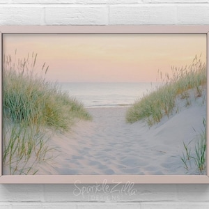 Peut inclure: Une photographie encadrée d'un chemin menant à l'océan à travers des dunes de hautes herbes. Le ciel est d'un rose et d'un orange doux au coucher du soleil.