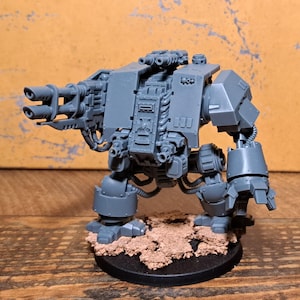 Op de afbeelding: Een grijze plastic miniatuur robotfiguur met een zwaar dubbelloops wapen aan de rechterarm en een mechanische klauw aan de linkerarm. Het figuur staat op een ronde zwarte voet met getextureerde bruine terreindetails op het oppervlak.