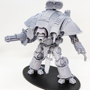Pode incluir: Uma miniatura de resina cinzenta sem pintura de um robô mecânico bípede. A figura apresenta um grande lançador de mísseis montado no ombro, um canhão gatling de vários canos no braço direito, uma mão com garras à esquerda e blindagem ornamentada.