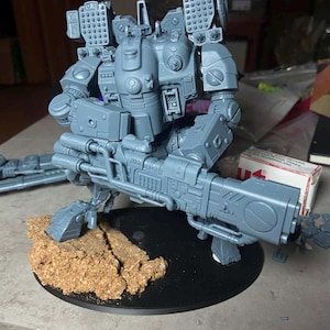 Op de afbeelding: Een grijs plastic model van een futuristische tweebenige robot op een zwarte ronde voet met een gestructureerd bruin terreinstuk. De robot is voorzien van op de schouder gemonteerde raketwerpers, een armkanon en gedetailleerde mechanische beplating.