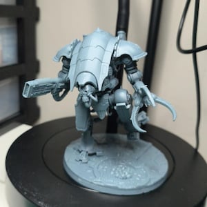 Op de afbeelding: Een grijze plastic miniatuur robotfiguur met een grote gebogen carapax, een mechanische arm met een meervoudig wapen en een klauwhand. Het figuur staat op een ronde basis, geplaatst naast een groene liniaal met metingen in millimeters.