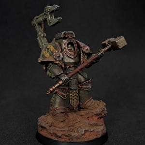 Op de afbeelding: Een gepantserd miniatuurfiguur in verweerde metallic groene en bronzen beplating, met een op de schouder gemonteerde mechanische klauw en een grote rechthoekige hamer. Het figuur staat op een getextureerde, roodbruine rotsachtige basis.