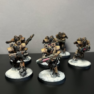 Op de afbeelding: Een groep van vijf miniatuur robotfiguren geschilderd in tinten bruin en beige met zwarte accenten. Elk figuur staat op een ronde witte basis en is uitgerust met een wapen, voorzien van metalen details en gloeiende roze energiecomponenten.