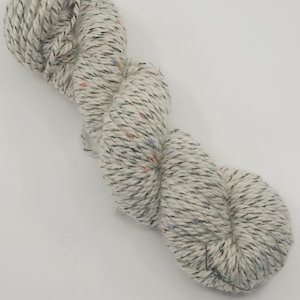May include: A skein of speckled yarn in a light gray color. The yarn has flecks of black, orange, blue, and green throughout. The yarn is twisted and appears soft, suitable for knitting or crocheting projects.